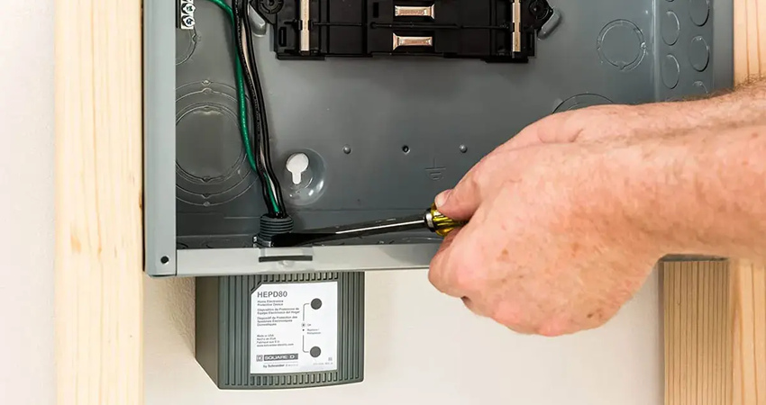 An electrician installs a whole home surge protector beneath a residential electrical panel.