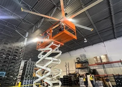 Commercial electrician installing large industrial ceiling fan in warehouse using scissor lift"